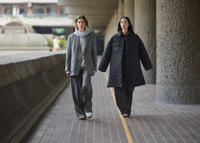 Два человека идут рядом под бетонными колоннами в одежде стиля Uniqlo: один в сером пиджаке и широких брюках, другой в длинном черном стеганном пальто